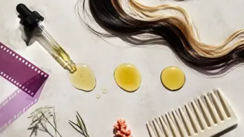 Tre gyldne oljedråper, hårstrå, pipette og kam på en lys mineralflate